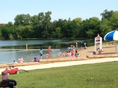 some people are sitting on the grass by the water and playing in the sand at the park