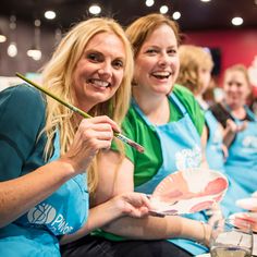 three women in aprons are smiling and holding paintbrushes while sitting at a table