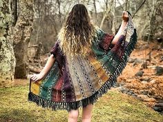 a woman standing in the woods with her back to the camera, wearing a multicolored shawl