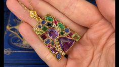 This gorgeous 18K yellow gold enhancer pendant is bezel set with six (6) natural blue sapphires, two (2) natural peridot, two (2) natural emeralds, two (2) natural jadeite jade, two (2) natural pink sapphires, two (2) natural purple sapphires, two (2) natural amethyst and eleven (11) round brilliant cut diamonds. The tapered bail is bead set with two (2) round brilliant cut diamonds. The pendant measures 48.00mm X 31.00mm and is suspended from a 14K yellow gold neck chain measuring 18 inches in length. One of the peridots is lightly chipped. Yellow Gold Multi-stone Art Deco Jewelry, Art Deco Multi-stone Yellow Gold Jewelry, Art Deco Yellow Gold Multi-stone Jewelry, Art Deco Yellow Gold Jewelry With Gemstone Accents, Gold Rectangular Multi-stone Jewelry, Fine Jewelry Cabochon Gemstone Pendant, Elegant Rectangular Multi-stone Jewelry, Elegant Multicolor Pendant Gemstones, Elegant Multicolor Gemstone Pendant