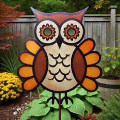 an orange and brown owl statue sitting on top of a lush green plant next to a wooden fence