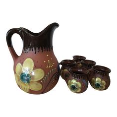 a brown pitcher with yellow flowers on it and six smaller cups in the foreground