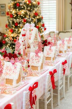 there are many small gingerbread houses on the table with red ribbon around them and a christmas tree in the background