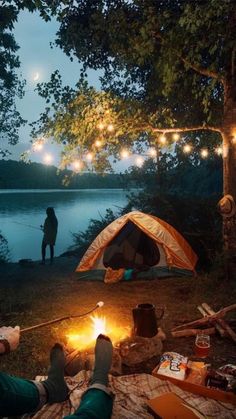 two people are camping in the woods at night with their tent and campfire lit by string lights