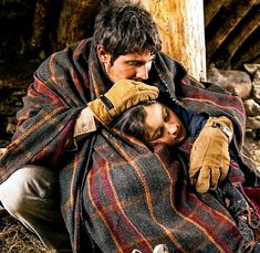 a man and woman wrapped in blankets sitting on the ground next to a wooden structure