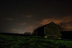 an old barn sits in the middle of a field at night with stars above it