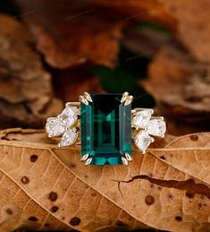 an emerald and diamond ring sitting on top of a leaf covered ground with leaves around it