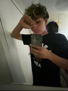 a young boy is looking at his cell phone in the mirror while wearing a black t - shirt