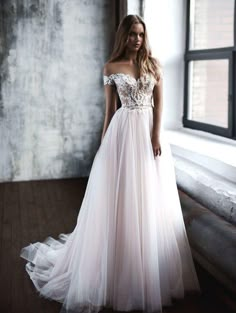 a woman standing in front of a window wearing a wedding dress with an off the shoulder design