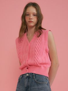 a woman in a pink sweater and jean skirt stands against a pink wall with her hands on her hips