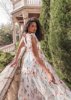 a woman in a white floral dress leaning on a stone wall and looking off into the distance