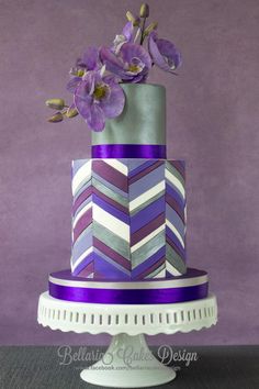 a three tiered cake with purple flowers on the top and silver chevron design