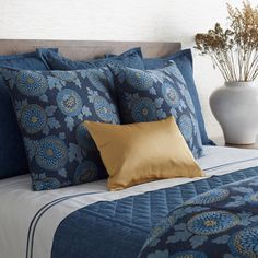 a bed with blue and gold pillows on top of it next to a white vase