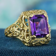 In brilliant yellow gold, this vintage-inspired ring showcases a wide filigree band that elegantly tapers toward the front. Intricate scrollwork filigree graces the band's shoulders, accentuated by a delicate milgrain touch for added detail. The focal point of the ring is a stunning 3.5 carat rectangular purple amethyst, featuring a dazzling faceted emerald-cut securely set within a bezel setting. With its captivating design and undeniable elegance, this ring is guaranteed to captivate attention and inspire admiration wherever you wear it. CHARACTERISTICS Status: Made to order Origin: Thailand Metal: Solid 9K Yellow Gold Ring Size: US 3-8 Total Gemstones Weight: 4.00 carat. (approx.) Total Gram Weight: 4.63 g. (approx.) *Pictures have been enlarged to show details* PRIMARY STONE(S) Stone: Luxury Victorian Amethyst Ring In Yellow Gold, Elegant Wide Band Gemstone Jewelry, Formal Wide Band Gemstone Ring, Wide Band Gemstone Rings For Formal Occasions, Ornate Yellow Gold Gemstone Rings, Ornate Engraved Yellow Gold Filigree Ring, Yellow Gold Gemstone Filigree Ring, Yellow Gold Engraved Ring With Intricate Design For Anniversary, Elegant Wide Band Engraved Ring For Anniversary