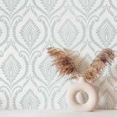 two vases with dried plants in front of a wallpapered background, one is white and the other is light blue