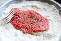 a metal bowl filled with flour and a red meat patty next to a fork in it