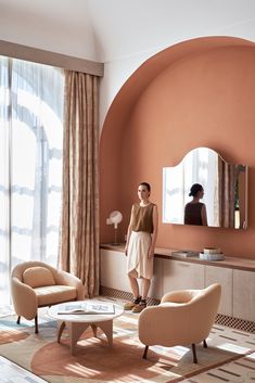 a woman standing in a living room next to two chairs and a table with a mirror on it