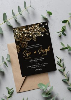 a black and gold wedding card with string lights on it next to some green leaves