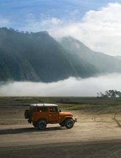 an orange jeep driving down a dirt road in front of a mountain covered with clouds