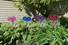 three colorful glass owls sitting on top of metal poles in front of plants and bushes