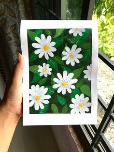 a hand holding up a card with white daisies on green leaves in front of a window