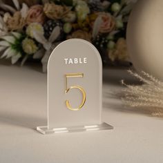 a white table number with gold numbers on it and flowers in the backgroud