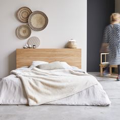 a woman walking past a bed in a room with white walls and wooden headboard