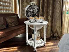 a white table with wine glasses on it in front of a brown couch and curtains