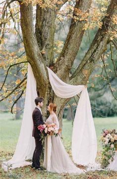 a bride and groom standing in front of a tree with white drapes on it