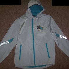 San Jose Sharks Nhl Hooded Jacket. New W/ Tags. The Jacket Has A Shark Skin Feel Polyester Outer Shell That Is Water Resistant. It's A Light To Medium-Weight Hooded Jacket With Full Zip Front.. The Inside Lining Is Fuzzy Felt Lining. The Hood Is Attached And Has A Draw String. The Jacket Has 2 Pockets On The Outside. The Sharks Logo On The Front Is Sewn On. Measurements Of The Jacket Side To Side Under Arms: S: 23: M: 24 L: 25" Xl: 26.5 2x: 28" The Sharks Lettering On The Sleeves Is Reflective. Casual Gray Outerwear For Winter Sports, Sporty Hooded Outerwear With Reflective Details, Gray Sporty Outerwear For Winter Sports, Sporty Gray Outerwear For Winter Sports, Gray Windproof Sports Outerwear, Gray Windproof Outerwear For Sports, Gray Sports Outerwear With Adjustable Hood, Reflective Long Sleeve Hooded Jacket For Outdoor, Outdoor Hooded Jacket With Reflective Details