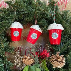 three red starbucks cups hanging from a christmas wreath with pine cones and evergreen branches around them