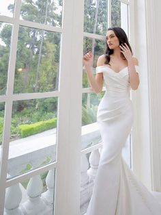 a woman in a white wedding dress standing by a window with her hand on her hip