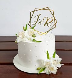 a wedding cake with white flowers and gold monogrammings on top, sitting on a wooden table