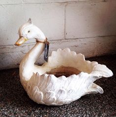 a white duck statue sitting on top of a floor next to a brick wall with a tag in it's beak