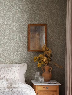 a vase with flowers on a table next to a bed in a room that has wallpaper