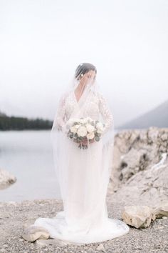 a woman in a white wedding dress standing on rocks near the water with her veil over her head