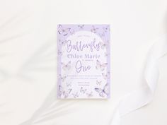 a purple and white wedding card with butterflies on the front, sitting next to a white ribbon