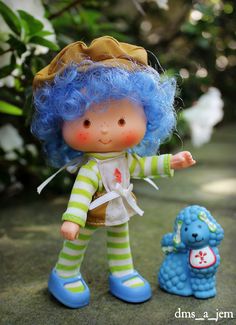 a blue haired doll standing next to a small toy bear on the ground with trees in the background