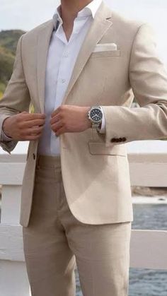 a man in a tan suit and white shirt is standing on a bridge by the water