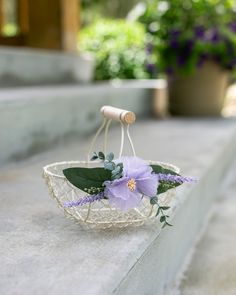 a white basket with purple flowers and greenery hanging from it's side on concrete steps