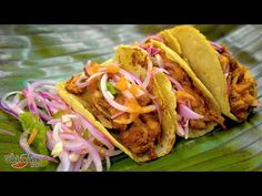 three tacos on a plate with onions and cheese in the middle, sitting on a leafy green surface