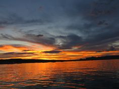 the sun is setting over water with clouds in the sky and on land behind it