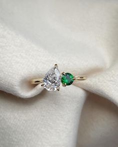 an engagement ring with two pear shaped diamonds and a green diamond on top, sitting on a white cloth