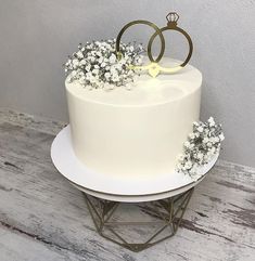 a white wedding cake with flowers and two gold rings on top, sitting on a metal stand