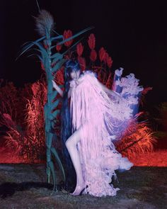 a woman in a dress standing next to a tall plant with feathers on it's head