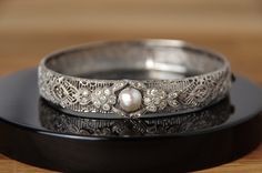 This is a stunning art deco style silver filigree clasp bracelet bangle, with cultured pearl (not tested) and with quartz or paste diamonds, engraved STERLING on the inside. One stone is missing from the front setting and there is some wear and slight bend to the shape of the bangle, please review photos thoroughly to see condition! The clasp with slide opening is in good working order and the filigree is in good condition.  Band width: 10mm Internal circumference: approx. 17.75cm All informatio White Gold Filigree Bracelet For Weddings, Elegant Engraved Bangle, Vintage Diamond Accented Bangle Bracelets, Vintage Bangle Bracelets With Diamond Accents, Elegant Engraved Jewelry For Weddings, Art Deco Silver Bracelets With Diamond Accents, Silver Filigree Bracelets For Wedding, Victorian Diamond Wedding Bracelets, Vintage Formal Bangle Hand Set