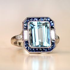 A stunning gemstone ring that centers on a 3.20 carat natural emerald-cut aquamarine. Baguette-cut diamonds and natural French-cut sapphires surround the aquamarine. Additional diamonds line the shoulders. This ring is platinum and displays an open-work under-gallery. ✦ GEMSTONE SPECIFICATIONS: Center Stone: Aquamarine Gemstone Weight: 3.20 Carats Gemstone Cut: Emerald Cut ✦ ENGAGEMENT RING SPECIFICATIONS: Stones: Aquamarine, Diamonds, Sapphires Material: Platinum Ring Style: Art Deco ✦ WHAT COM Diamond Ring With Halo, Emerald Cut Aquamarine Ring, Sapphire Engagement Ring Halo, Ring With Halo, Estate Diamond Jewelry, Emerald Cut Rings, Gemstone Engagement, Baguette Cut Diamond, Aquamarine Rings