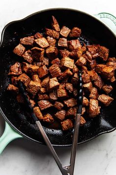 a skillet filled with cooked meat sitting on top of a counter next to two tongs