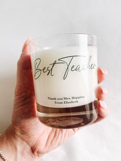 a person holding a glass with the words best teacher on it in front of a white background