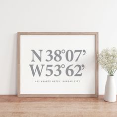 a white vase sitting next to a framed poster on a wooden table with a plant in it